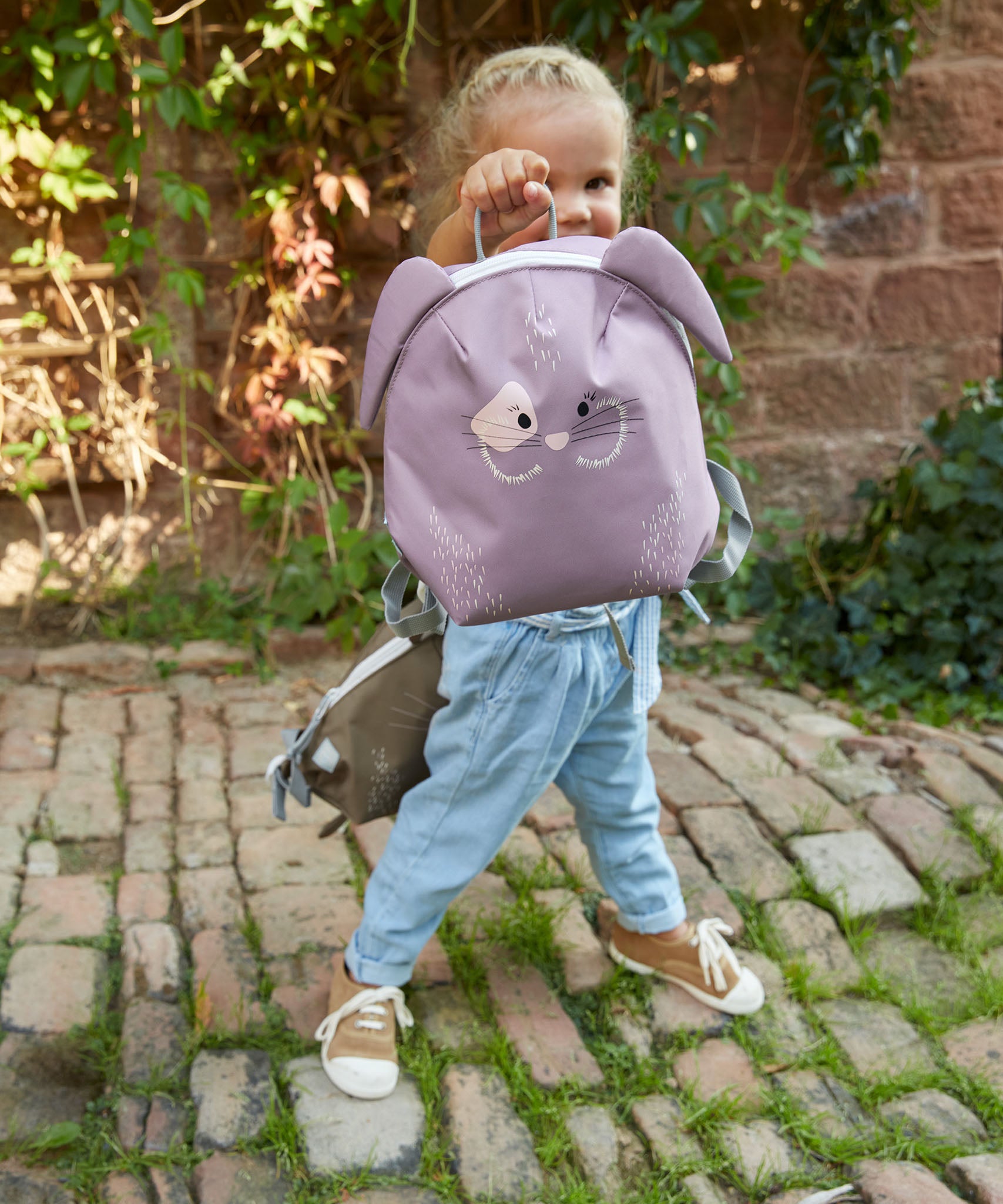 A child standing on a cobbled path and holding the Lassig bunny tiny backpack by the loop handle on the top