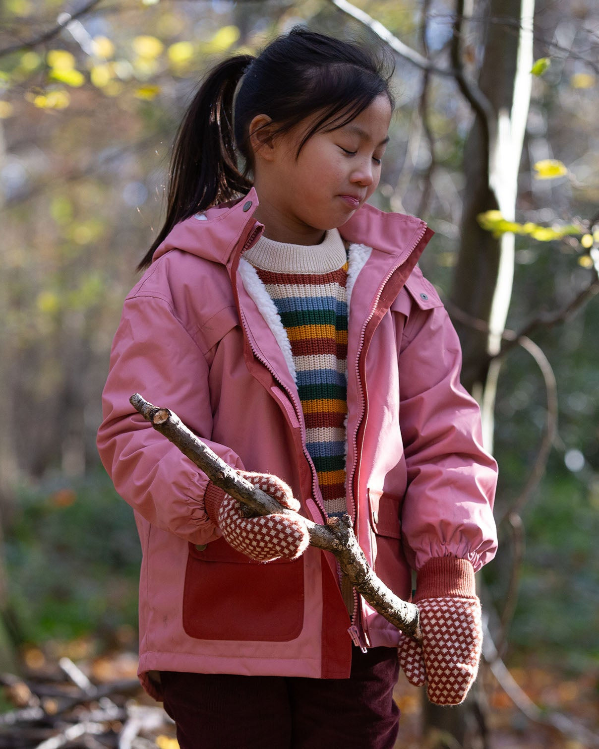 child wearing pink little green radicals coat holding a branch in a forest setting