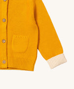 A close up of the Little Green Radicals gold snuggly hooded knitted cardigan. Showing the knitted pocket and cream cuff detail on yellow organic cotton fabric.