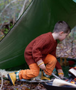 A child wearing the Little Green Radicals ginger aran knitted jumper in woodland under a shelter. A thick cable knit style jumper in dark orange GOTS organic cotton fabric with striped cuffs.
