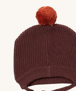 A close up of the Little Green Radicals chocolate knitted beanie hat showing the knit detail and light brown pom pom on stitched brown organic cotton fabric.