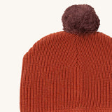 A close up of the Little Green Radicals ginger knitted beanie hat showing the knitted detail and purple pom pom on dark orange organic cotton fabric.