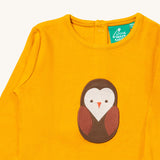 A close up of the Little Green Radicals gold brown owl applique long sleeve top showing the applique and button fastener detail. Features a small mushroom applique on the sleeve on dark yellow organic cotton fabric