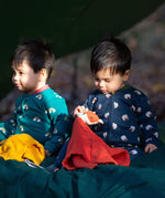 A child sitting on the floor in the woods and wearing the Little Green radicals navy sheep baby grow and holding a fox comforter. A soft GOTS organic cotton all in one in navy with cute sheep prints