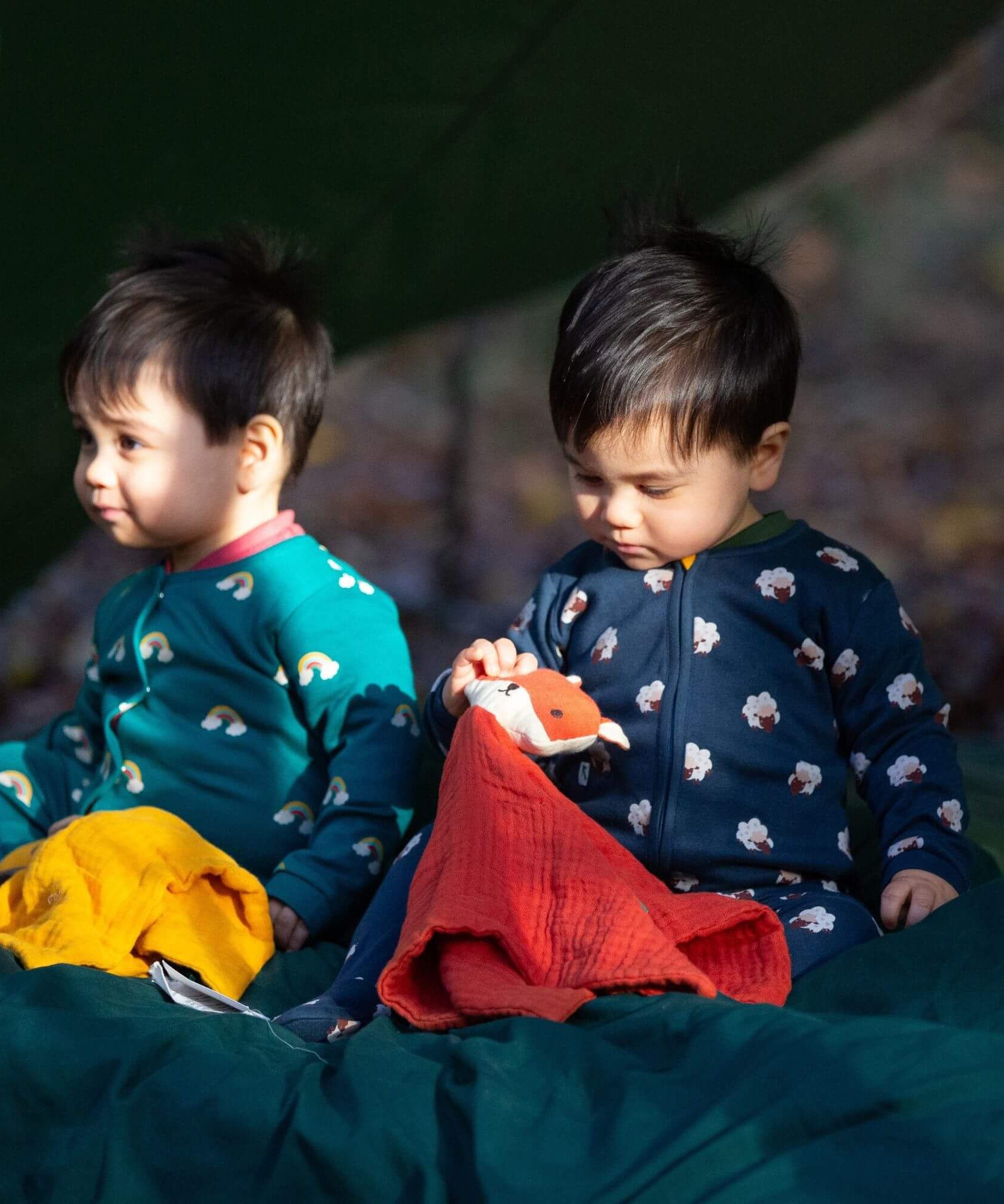 A child sitting on the floor in the woods and wearing the Little Green radicals navy sheep baby grow and holding a fox comforter. A soft GOTS organic cotton all in one in navy with cute sheep prints