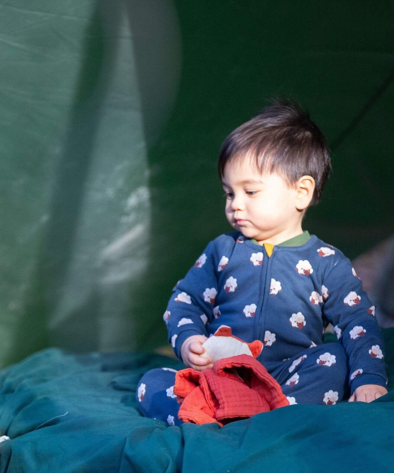 A child sitting under a shelter outside and wearing the Little Green radicals navy sheep baby grow. A soft GOTS organic cotton all in one in navy with cute sheep prints. Features a full length zip fastener with a yellow toggle