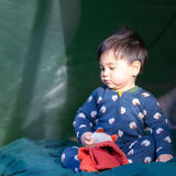 A child sitting under a shelter outside and wearing the Little Green radicals navy sheep baby grow. A soft GOTS organic cotton all in one in navy with cute sheep prints. Features a full length zip fastener with a yellow toggle