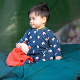 A child sitting on a green blanket under a shelter in the woods and wearing the Little Green radicals turquoise rainbow baby grow. A soft GOTS organic cotton all in one in navy with long sleeves and cute sheep prints