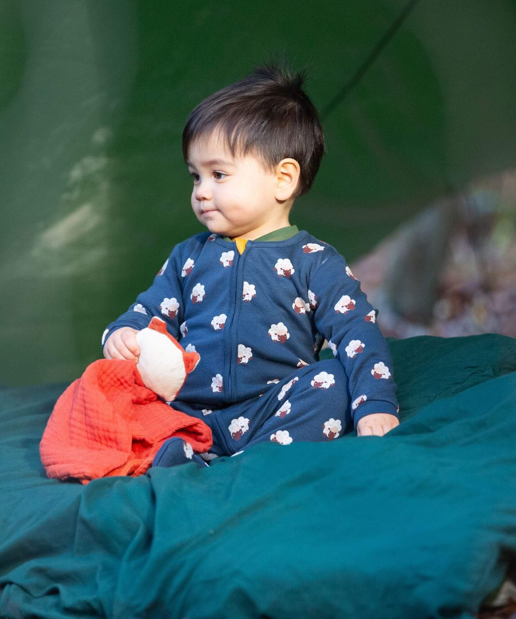 A child sitting on a green blanket under a shelter in the woods and wearing the Little Green radicals turquoise rainbow baby grow. A soft GOTS organic cotton all in one in navy with long sleeves and cute sheep prints
