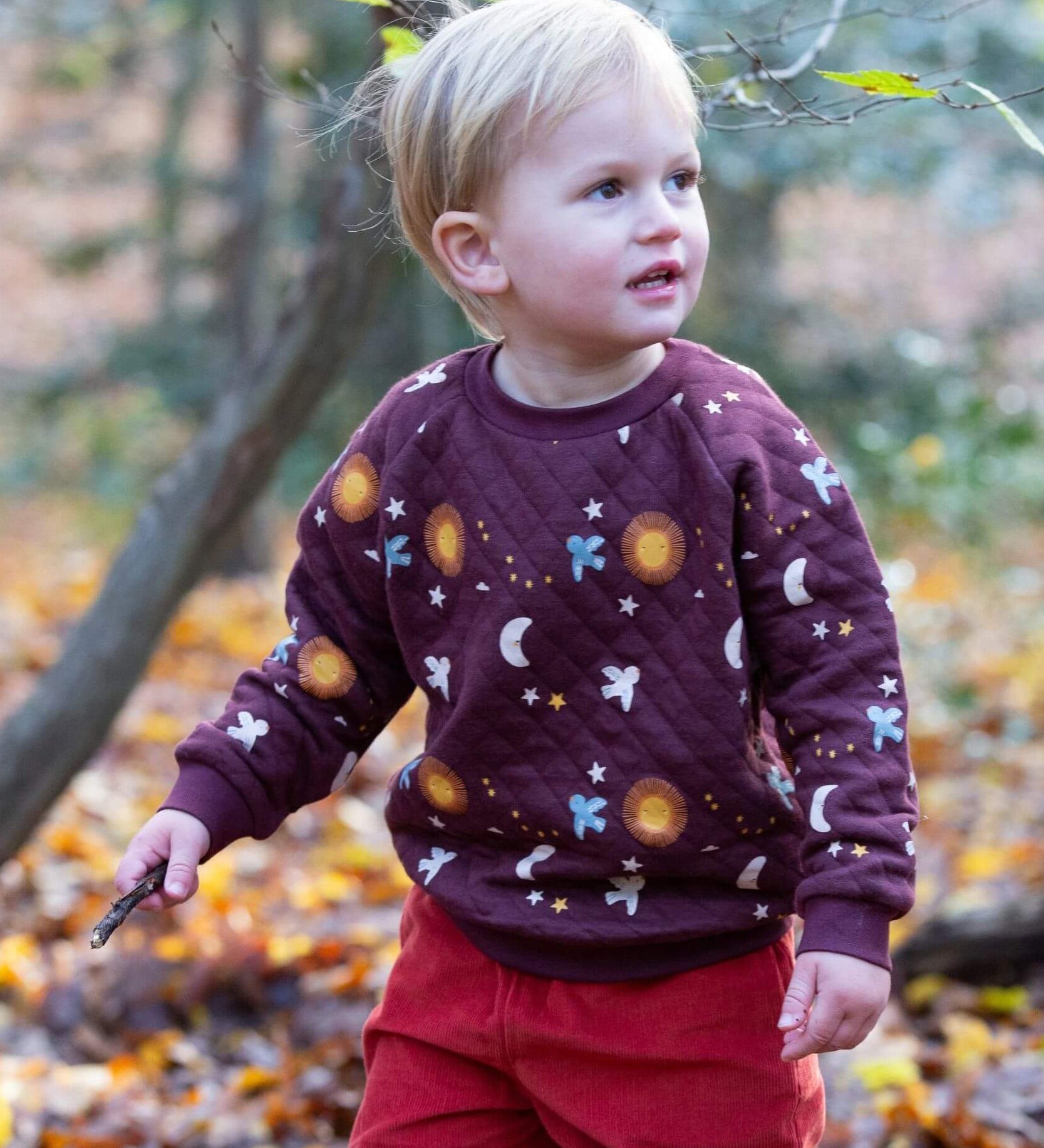 A child playing in woodland and wearing the Little Green Radicals flying birds quilted sweatshirt that's available at Babipur. A thick quilted sweatshirt with cute prints of birds flying through a starry night on dark maroon organic cotton fabric.