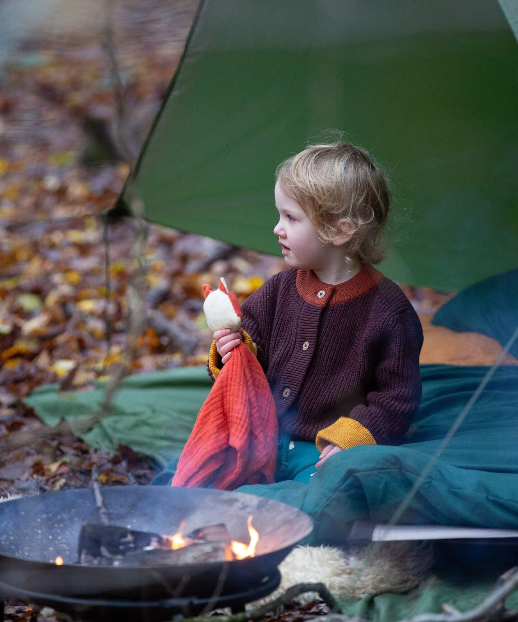 A child sitting outside and wearing the Little Green Radicals knitted cardigan in chocolate available at Babipur. A thick warm brown GOTS organic cotton cardigan with yellow arm cuffs, light brown collar and button fasteners.