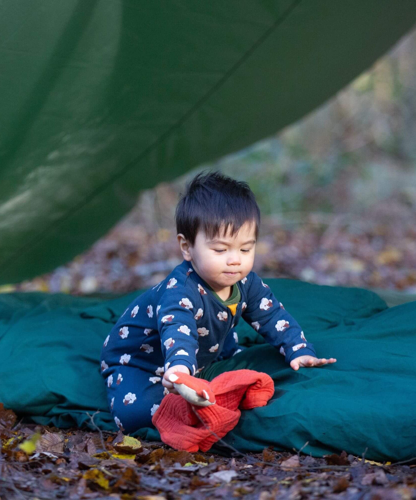 A child sitting on a green blanket and wearing the Little Green radicals turquoise rainbow baby grow with a sheep design, available at Babipur.