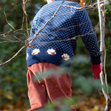A child standing in woodland and wearing the Little Green Radicals navy sheep snuggle knitted jumper. Showing the white speckles and cute sheep continuing around the jumper