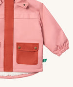A close up of the Little Green Radicals waterproof coat in coral Showing the elasticated cuff detail and dark pink side pocket on pink recycled polyester fabric
