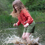 A child wearing a Little Green Radicals From One To Another Burnt Ochre Pointelle Knitted Cardigan with green coloured shorts. The child is standing in water, splashing. Little green Radicals have a wide range of organic cotton, Fairtrade children's clothing and outerwear available here at Babipur.