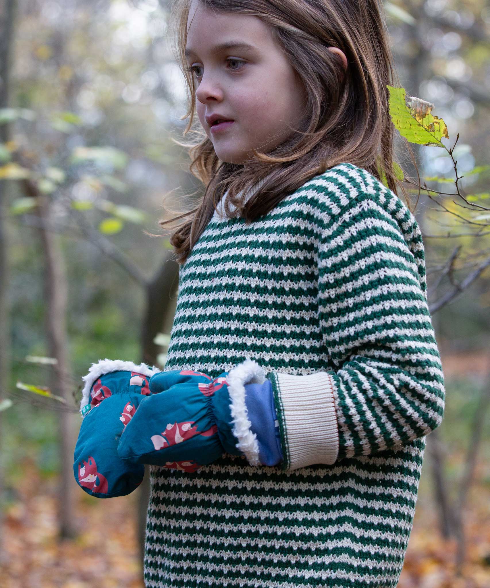 A child wearing the Little Green Radicals fox print sherpa fleece lined mittens with a olive green striped knitted jumper. The children's sized mittens are part of a wide range of organic cotton clothing for children available here at Babipur.