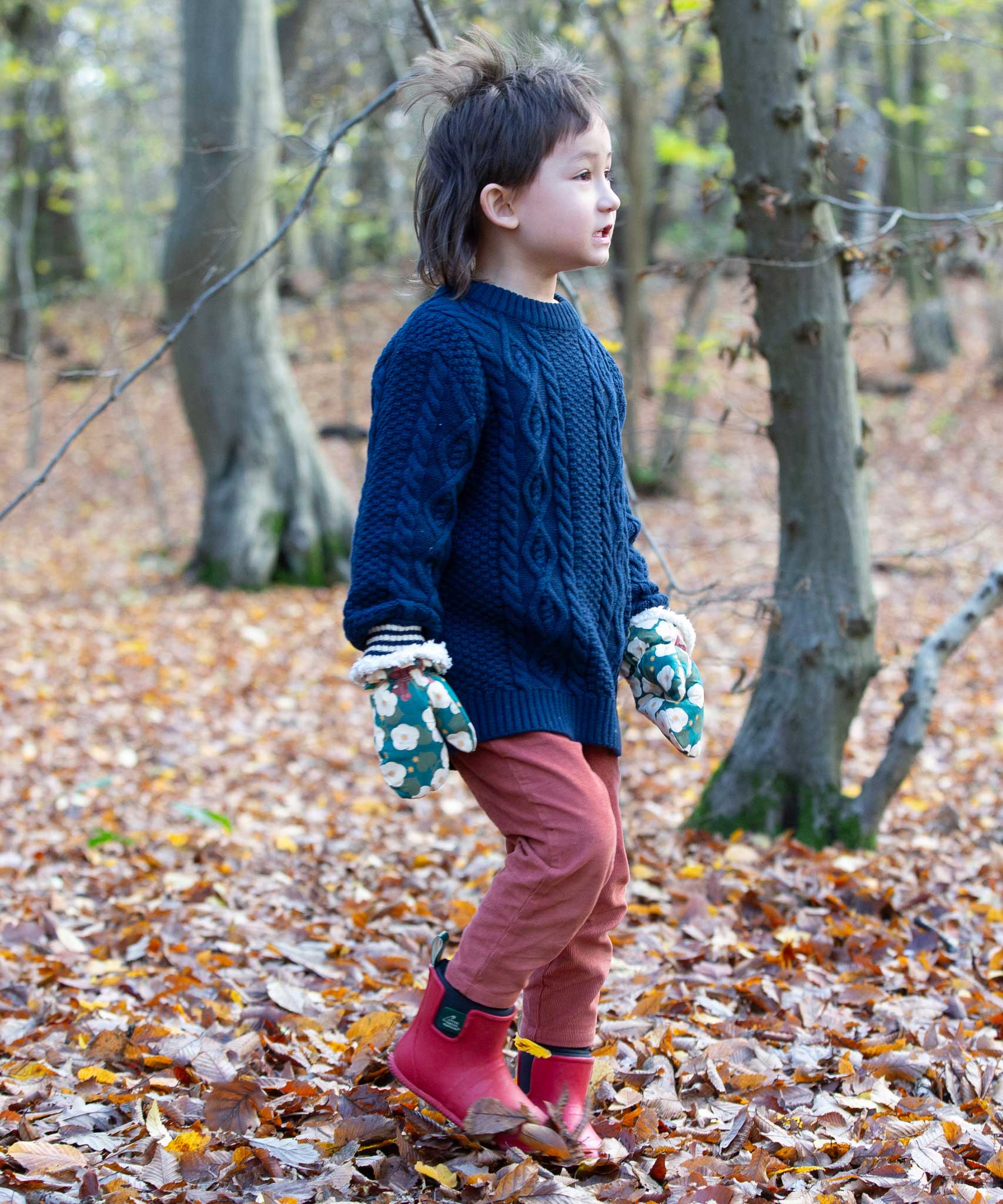 A child walking through a forest on a floor covered with fallen leaves wearing the  Little Green Radicals Hedgerow print Children's Sherpa Lined Mittens with a navy knitted jumper and ginger corduroy trousers.