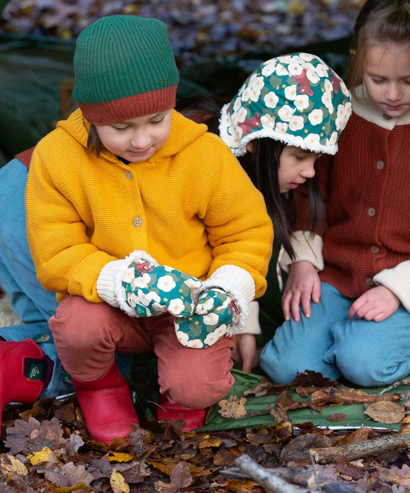 A child wearing the  Little Green Radicals Hedgerow print Children's Sherpa Lined Mittens with a yellow gold knitted cardigan and ginger orange corduroy trousers. There is a child in the background wearing a hedgerow sherpa lined hat.