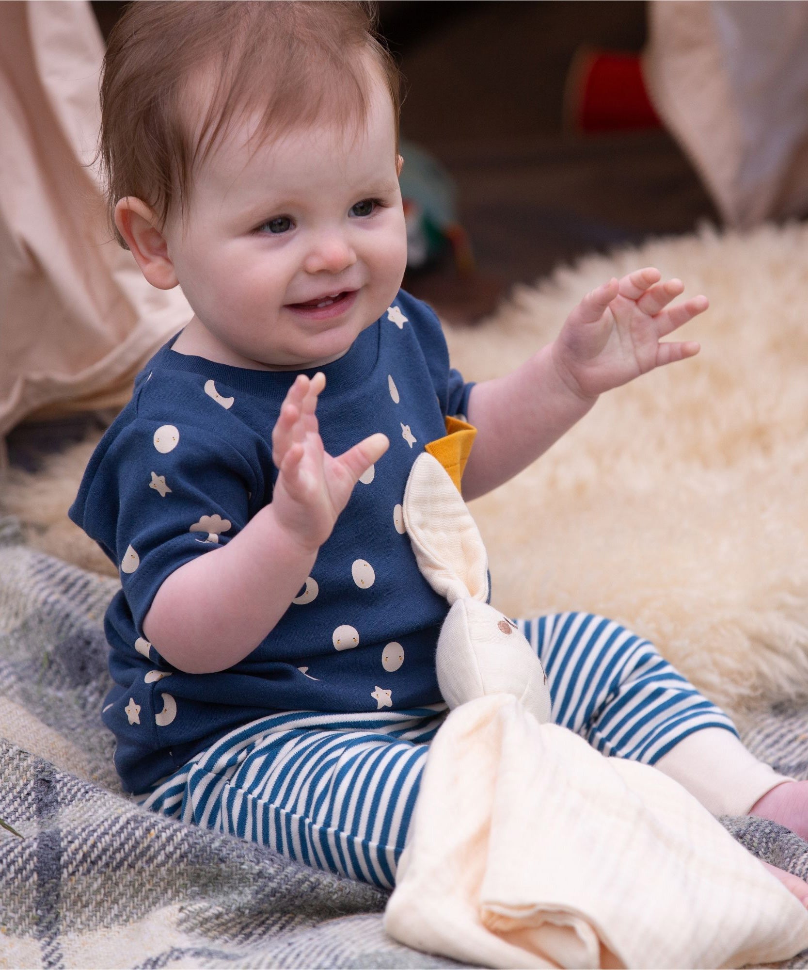 A baby wearing the Little Green Radicals  Full Moon T-Shirt & Jogger outfit set. The baby is sitting on a blanket with a cream coloured bunny comforter toy on their lap.