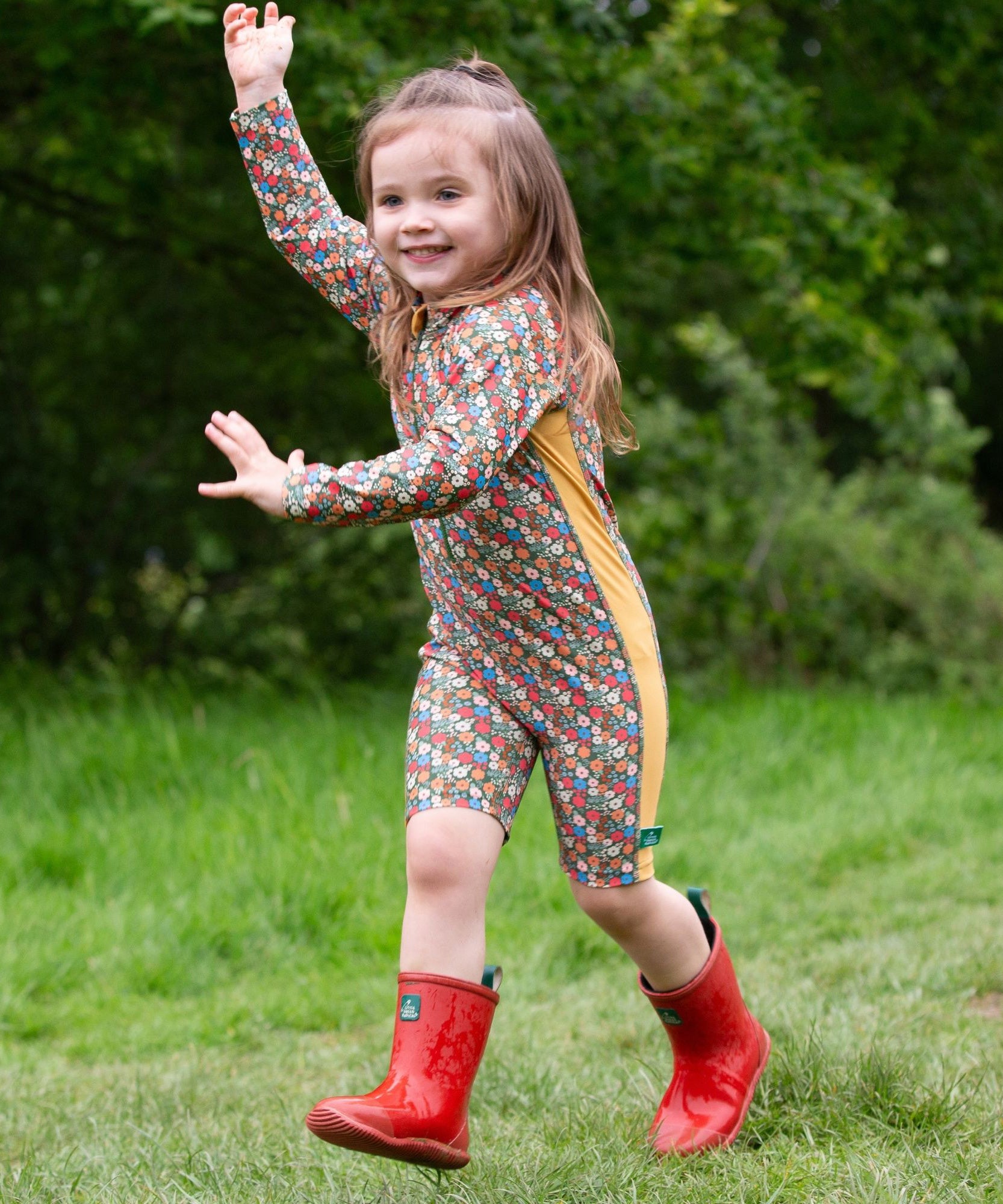 A child wearing the Little Green Radicals Vintage Flowers UPF 50+ Recycled Sunsafe Sunsuit with a pair of red wellington boots.