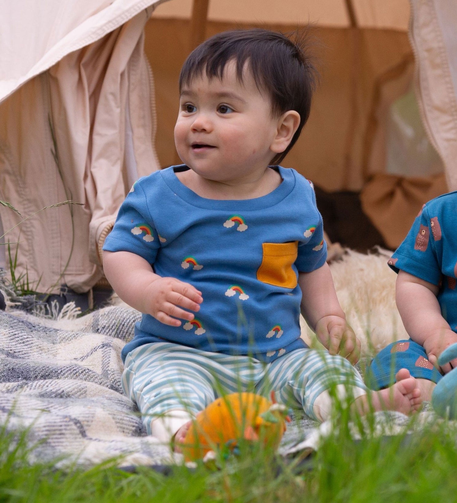 A young child sitting outdoors wearing the Little Green Radicals Kids' Organic T-shirt and Joggers Playset outfit in the  Blue Rainbows design.