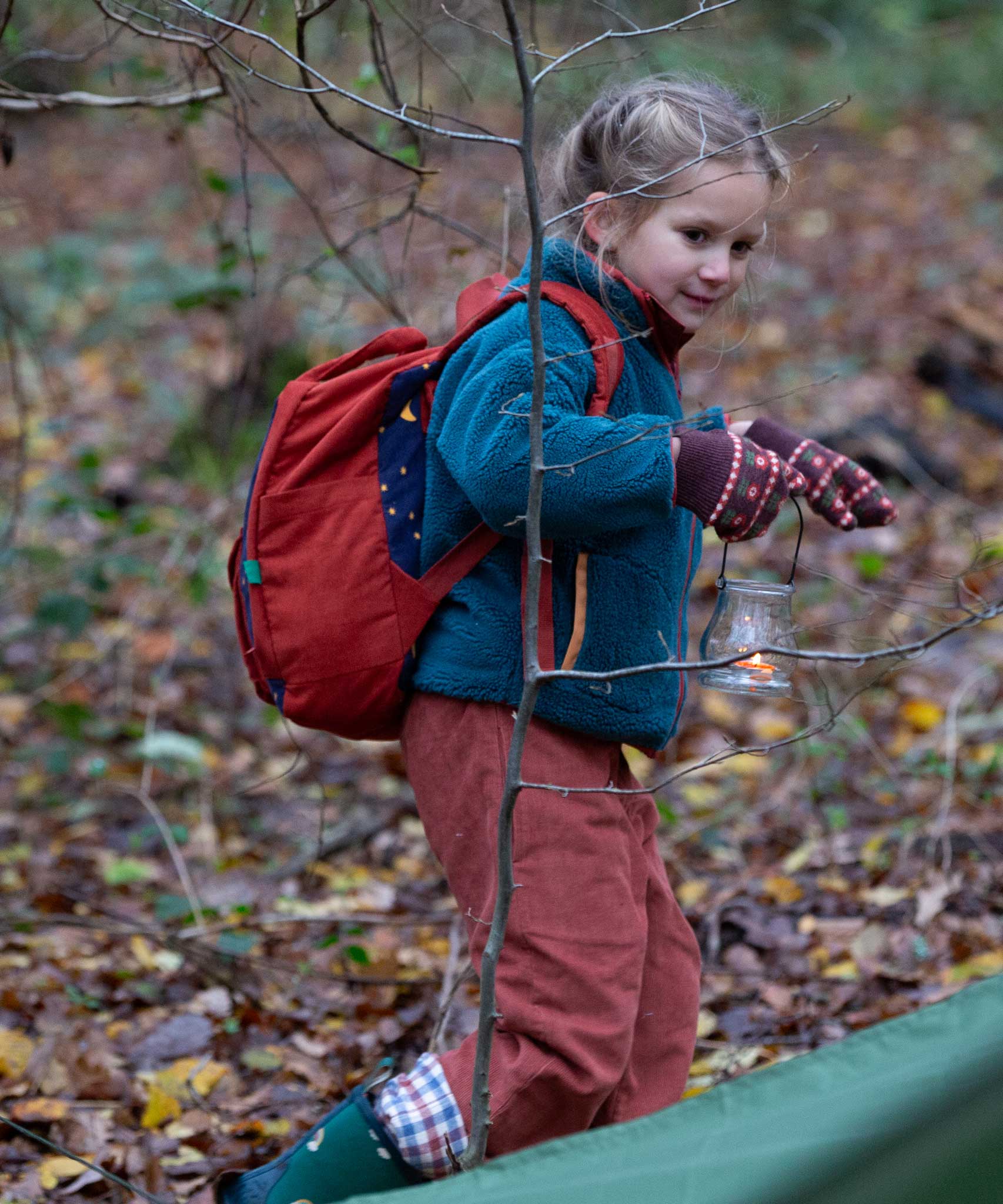 A child walking through a forest wearing the Little Green Radicals dark nights corduroy backpack on their back. A 100% organic cotton backpack in a navy and red corduroy style with a starry night design. Features a handle on the top for easy carrying a front pocket and 2 bottle holders. This children's bag is a part of a wide range of organic cotton clothing and accessories for children available here at Babipur.