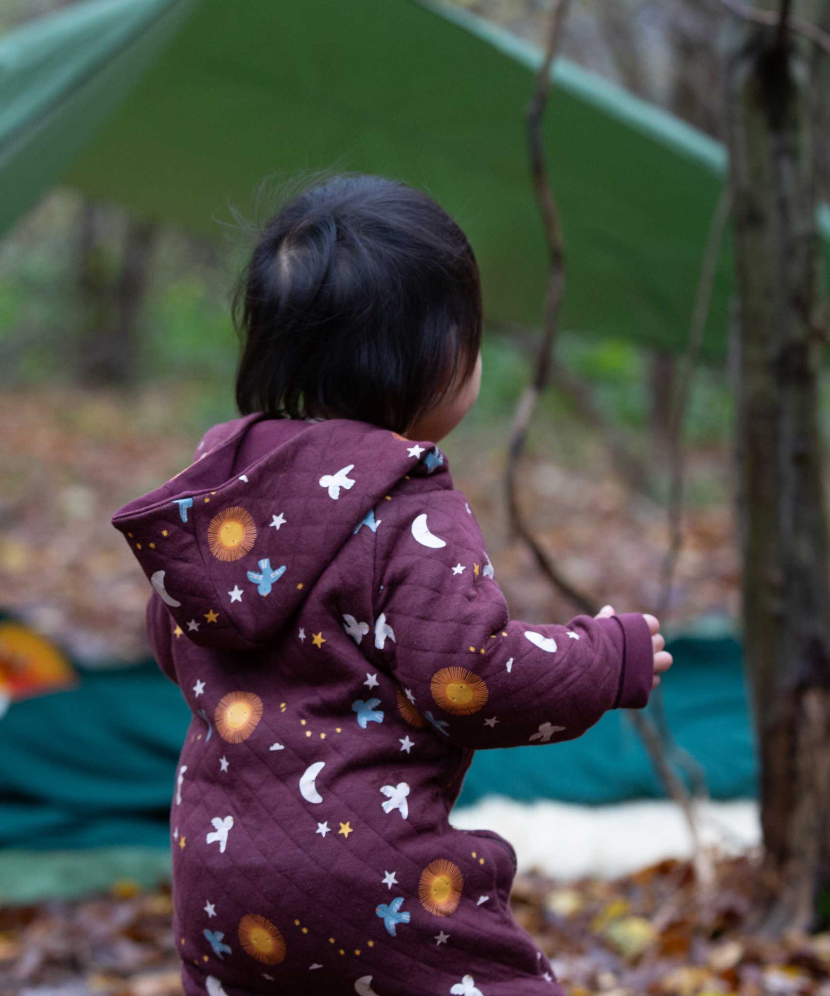 A view of the back of a child wearing the Little Green Radicals Organic Cotton Flying Birds Quilted Snug Suit. A maroon brown coloured suit with an all over print featuring flying birds, stars, moon and  sun. This snug suit is a part of a wide range of organic cotton clothing for children available here at Babipur.