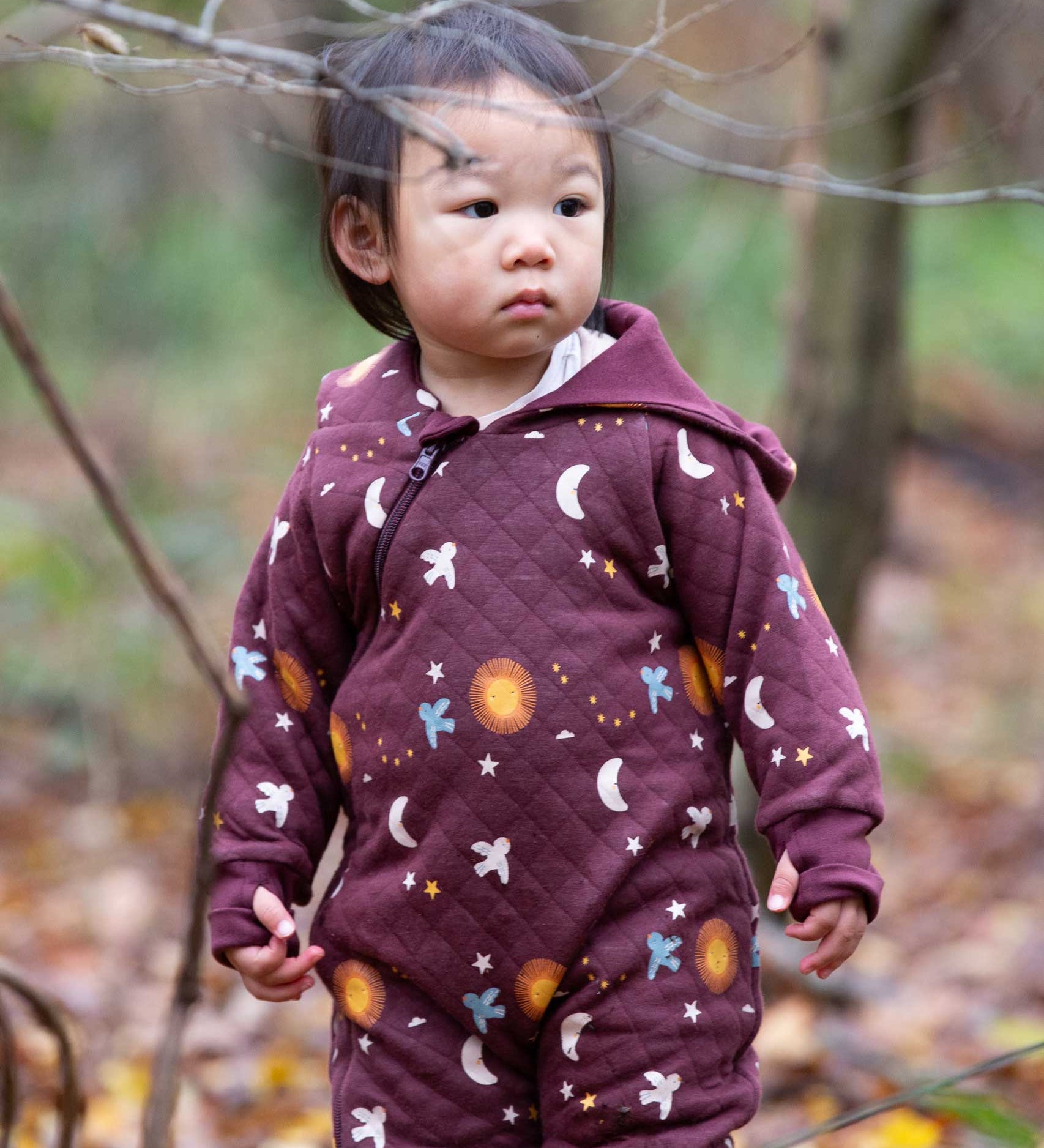 A child wearing the Little Green Radicals Organic Cotton Flying Birds Quilted Snug Suit. A maroon brown coloured suit with an all over print featuring flying birds, stars, moon and  sun. This snug suit is a part of a wide range of organic cotton clothing for children available here at Babipur.