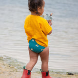 A young child wearing a Little Green Radicals Teal Rainbows UPF 50+ Recycled Reusable Swim Nappy with a yellow gold coloured t-shirt. The child is facing away from the camera.