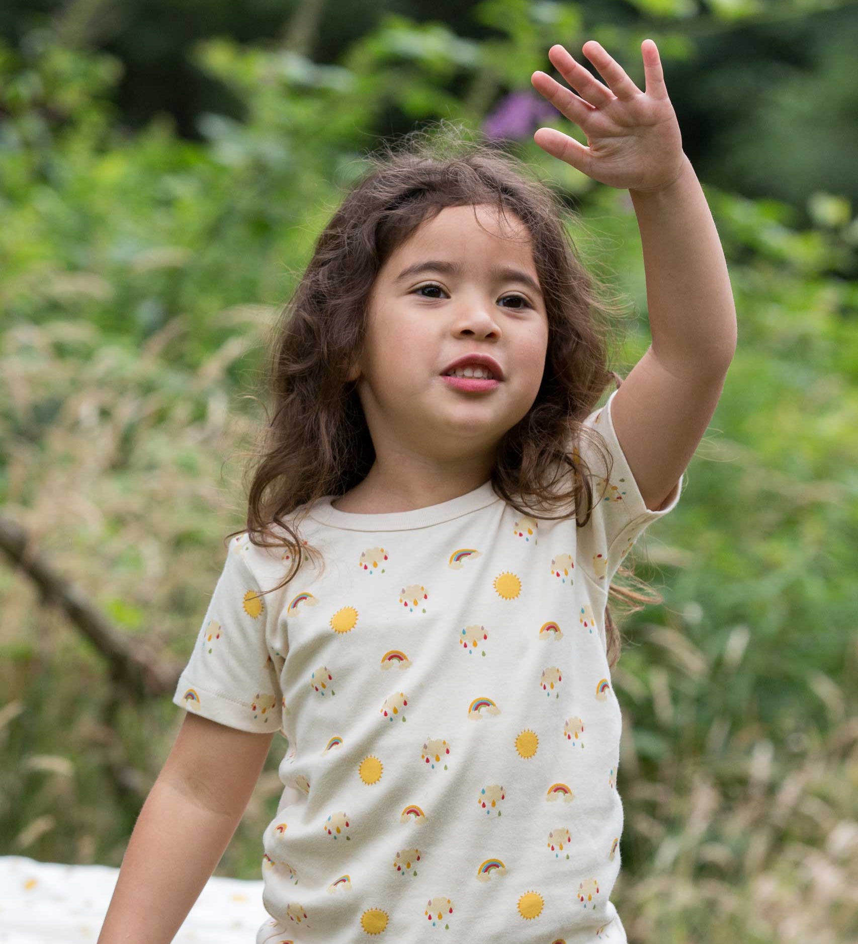 A child waving, wearing the Little Green Radicals Sunshine & Rainbows Short Sleeve organic cotton T-Shirt. An off white, cream coloured t-shirt with an all over print featuring rainbows, sunshine and rainbow rainclouds. Little green Radicals have a wide range of organic cotton, Fairtrade children's clothing and outerwear available here at Babipur.
