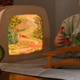 A Toverlux MagicLamp placed on a table with little red riding hood artwork placed inside. The lamp is on. There is a young child playing with an ostheimer little red riding hood wooden toy figure besides the lamp.