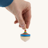 A wooden Mader traditional rainbow spinning top held by a hand on a cream background.