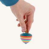 A wooden Mader dragon egg spinning top with multicoloured stripes  held by a hand on a cream background