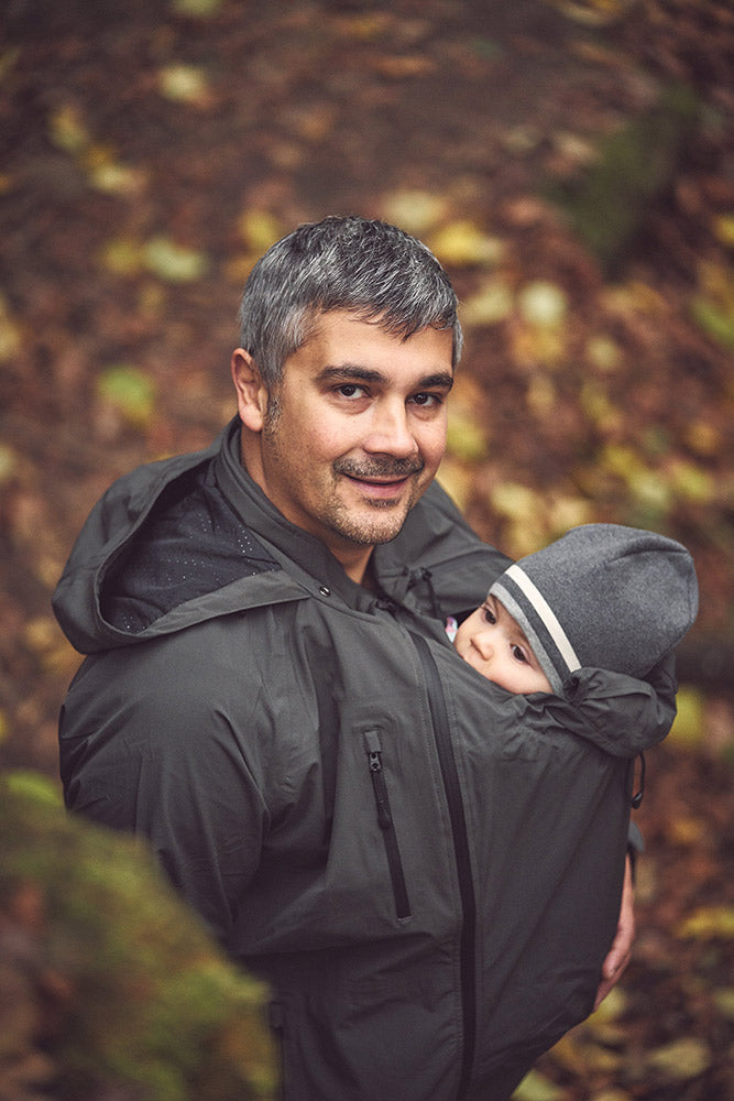Mamalila Shelter Men's Babywearing Rain Jacket in Anthracite. Lifestyle view of this men's technical babywearing rain coat with the baby carrier insert on the front. Woodland background