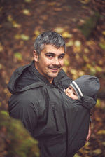 Mamalila Shelter Men's Babywearing Rain Jacket in Anthracite. Lifestyle view of this men's technical babywearing rain coat with the baby carrier insert on the front. Woodland background