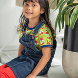 Young girl sat down wearing the Maxomorra eco-friendly navy denim pinafore dress over the short sleeve fox top