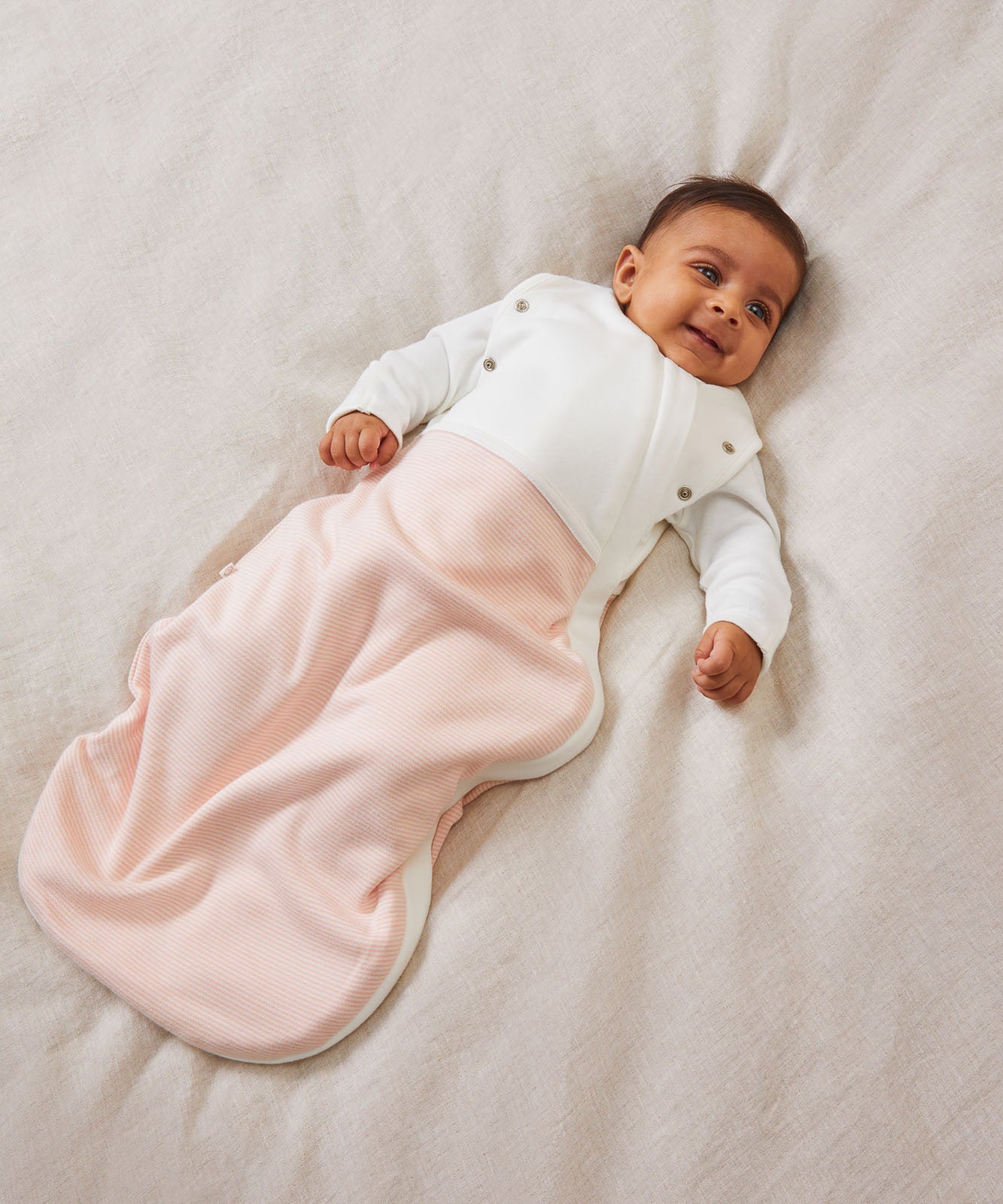 A baby laid on a soft surface inside the blush stripe MORI swaddle. Made from bamboo and organic cotton and features poppers on the arms, to tuck the arms in or let them be free.