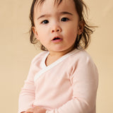A toddler sitting down, and wearing the Mori organic cotton and bamboo blend baby kimono bodysuit in a pink blush stripe design.