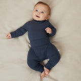 A baby laying on a bed, wearing a matching set of Mori navy comfy joggers made from organic cotton, and a long sleeve top.
