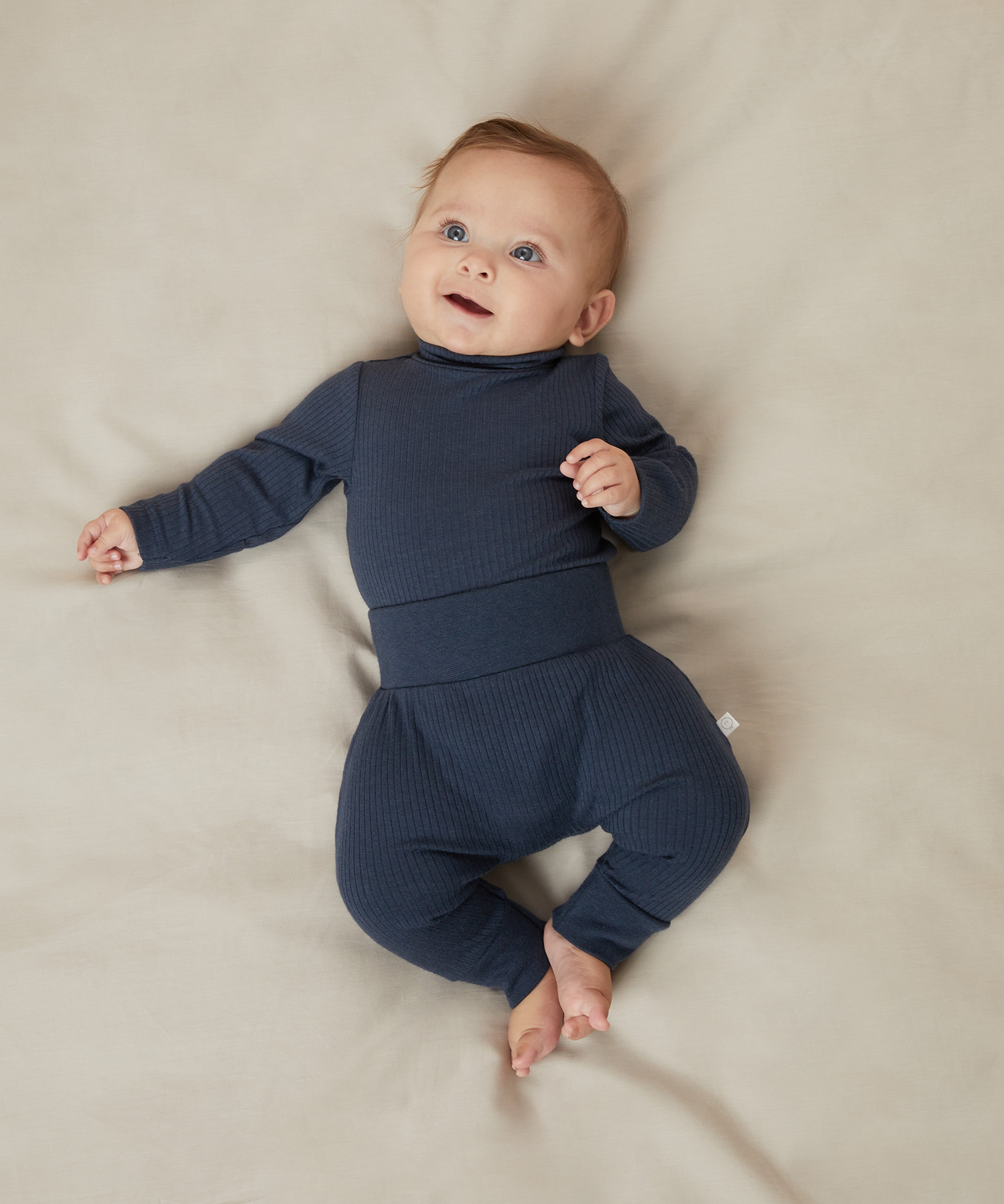 A baby laying on a bed, wearing a matching set of Mori navy comfy joggers made from organic cotton, and a long sleeve top.