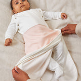 A baby laid on a soft surface inside the blush stripe MORI swaddle. Showing adults hand zipping the bottom half of the swaddle to tuck the baby in.