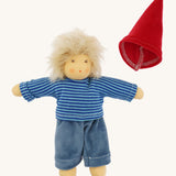 The Nanchen Frederik organic Waldorf doll dressed in a blue striped shirt and soft blue trousers, with the red gnome-style hat placed beside it. The doll’s blond mohair hair is fluffy and unstyled, and its hand-painted features are visible.