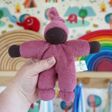 A Nanchen Pimpel Waldorf Handcrafted Doll with a berry pink coloured outfit and matching knotted hat being held up in an adult's hand in the Babipur playroom.