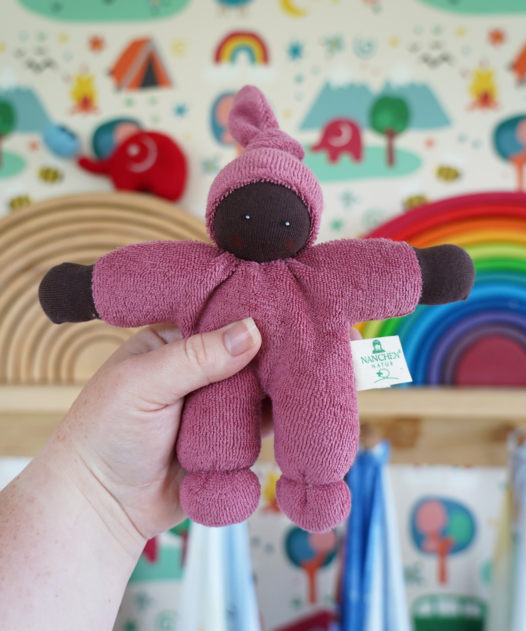 A Nanchen Pimpel Waldorf Handcrafted Doll with a berry pink coloured outfit and matching knotted hat being held up in an adult's hand in the Babipur playroom.