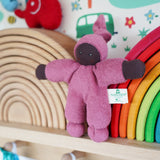 A Nanchen Pimpel Waldorf Handcrafted Doll with a berry pink coloured outfit an matching knotted hat. The doll has been placed on a wooden shelf in the Babipur playroom in front of two Grimm's wooden rainbow stacking toys, one is natural and one is rainbow coloured.