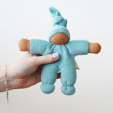 The Nanchen Pimpel Waldorf Doll in sage green is shown being held in an adult’s hand, highlighting its small, baby-friendly size. The doll’s face is hand-painted and it has a soft terry cloth body with a gentle expression.