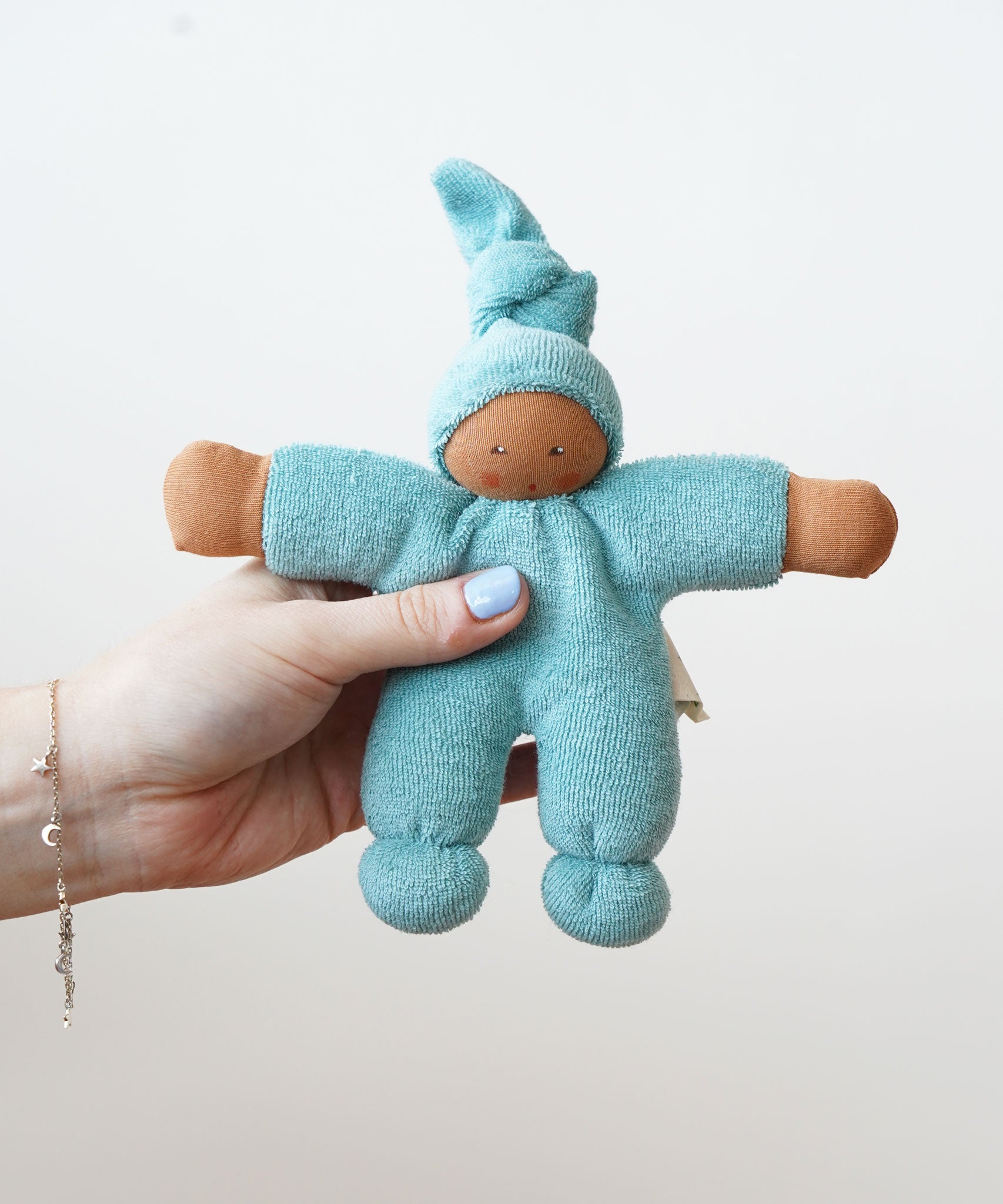 The Nanchen Pimpel Waldorf Doll in sage green is shown being held in an adult’s hand, highlighting its small, baby-friendly size. The doll’s face is hand-painted and it has a soft terry cloth body with a gentle expression.
