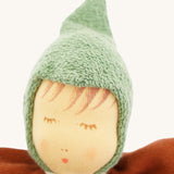 Close-up of the Nanchen Walnut Baby Doll's face, showing delicate hand-painted details including closed eyes with lashes, subtle blush, and soft lips. The doll wears a textured green hood made of organic cotton terry.