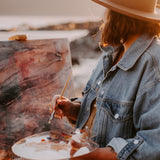 An artist outdoors holding a palette with Natural Earth Paint acrylics painting on a canvas