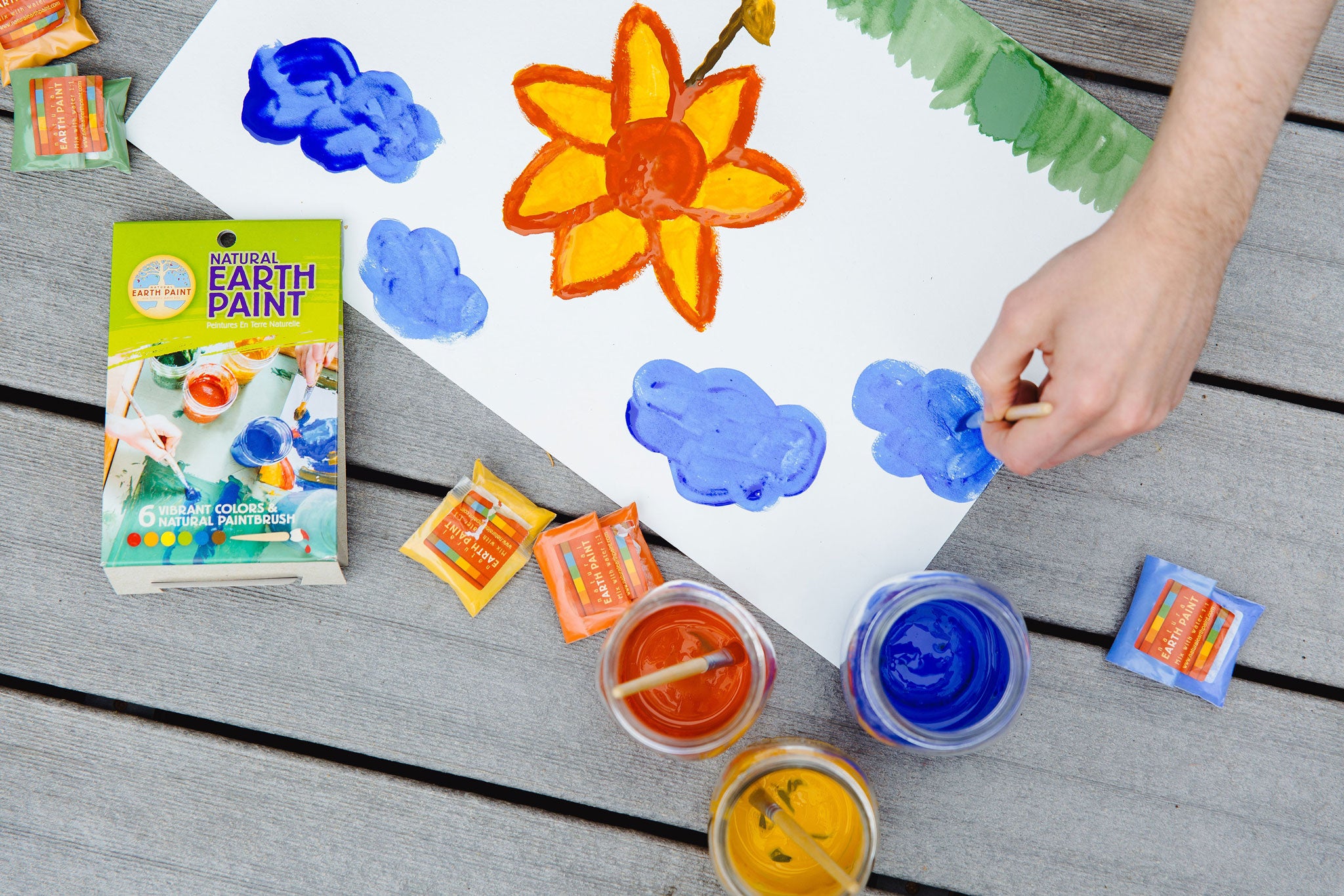 An overhead view of the Natural Earth Paint Petite Paint Kit in use. A child's hand can be seen holding the paintbrush, painting blue clouds.